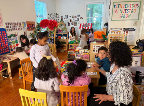 A colorful classroom features table groupings with preschoolers and educators gathered around, talking and smiling.