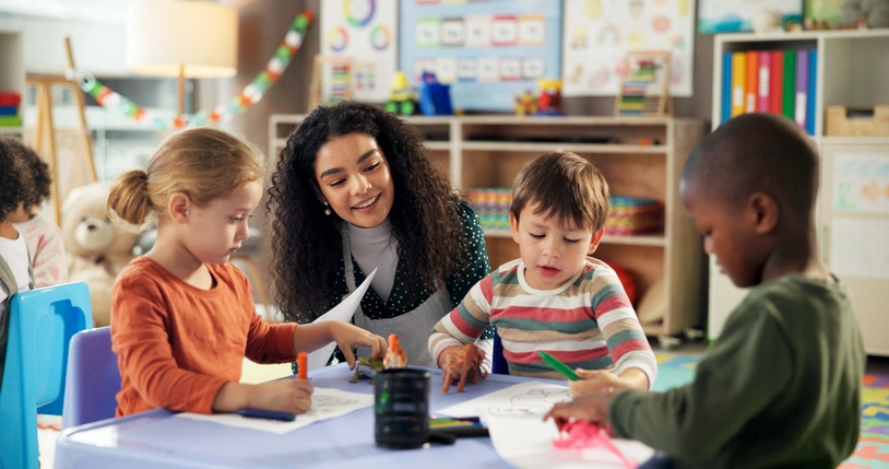 An educator and three young children sit around a table, coloring, in a preschool classroom.