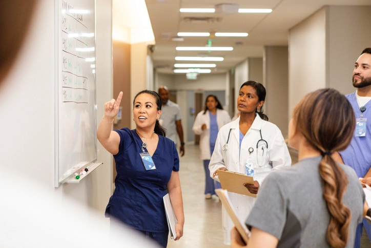 A nurse in scrubs points to a whiteboard on a wall, with doctors and nurses looking to the board.