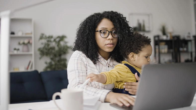 A woman in glasses sits at a table with a toddler on her lap, working on a laptop.