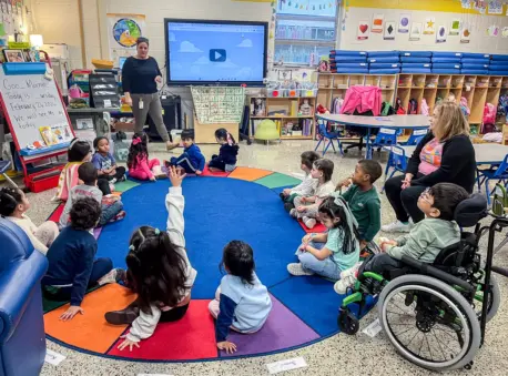 Young children sit in a circle around a blue rug in a classroom. A child raises her hand as a teacher looks on.